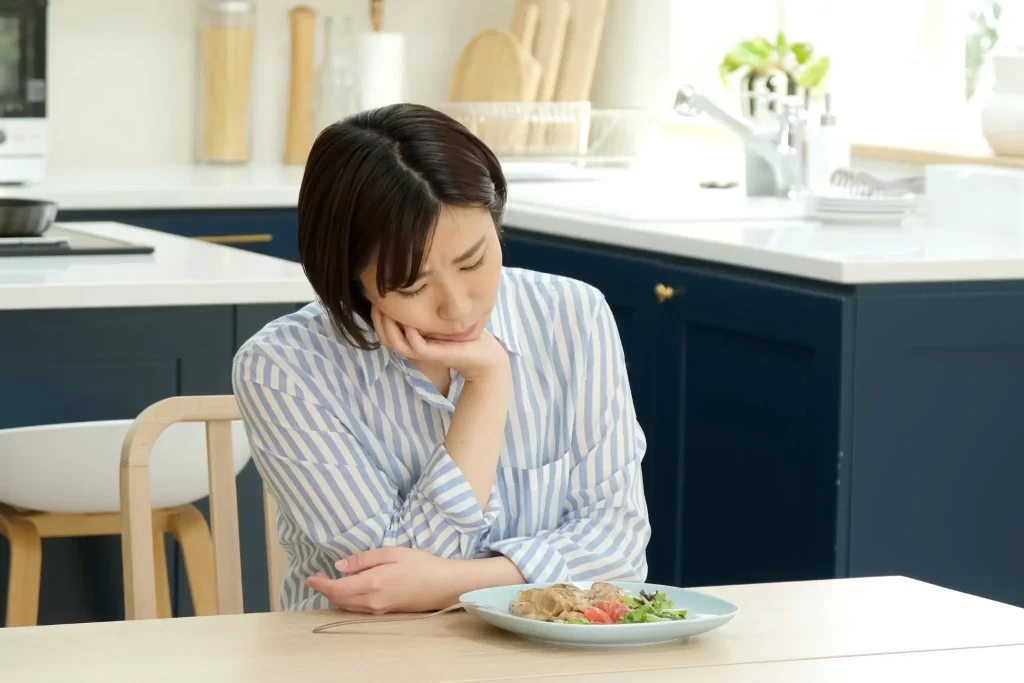 食事を前にうつむき悩む女性。ストレスによる食欲コントロールの難しさを表現。